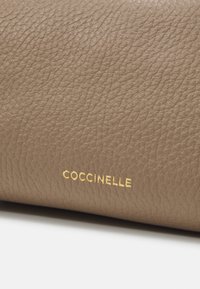 Tan krāsas ādas maciņš ar teksturētu virsmu un zeltītu "Coccinelle" logotipu iepresētu priekšpusē. Kompakts dizains daudzpusīgai lietošanai.