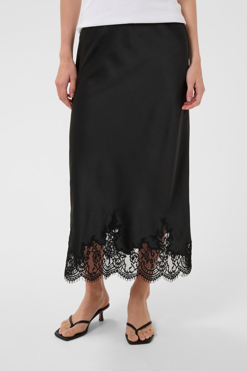 Femme portant une jupe midi noire avec un bord en dentelle et des sandales noires à talons, debout sur un fond blanc uni.