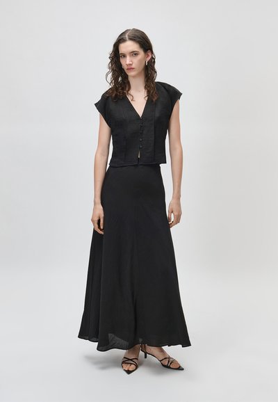 Femme aux cheveux bouclés portant un haut noir à manches courtes boutonné, une longue jupe noire et des chaussures à talons noires à brides sur un fond uni.