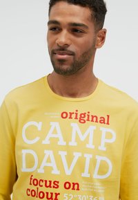 Junger Mann mit kurzen lockigen Haaren und Bart, trägt einen gelben Pullover mit großem weißen und roten CAMP DAVID Schriftzug.