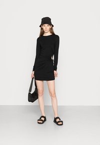 Vestido preto de manga longa com franzido lateral, combinado com um chapéu preto tipo balde e sandálias. Os acessórios incluem uma pequena bolsa preta de ombro.