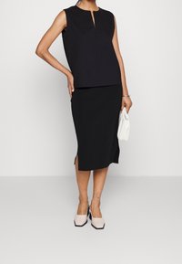 Black sleeveless top with a front keyhole cutout, paired with a black skirt featuring side slits. Model holds a small white handbag.