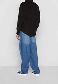 Camicia nera in corduroy, jeans larghi blu chiaro e comodi, e sneakers a fantasia blu e nere. Vista da dietro su uno sfondo bianco.