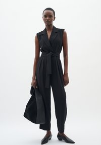 Salopette sans manches noire avec un design croisé et une taille ceinturée, assortie à un sac noir et des ballerines à bout pointu sur un fond clair.