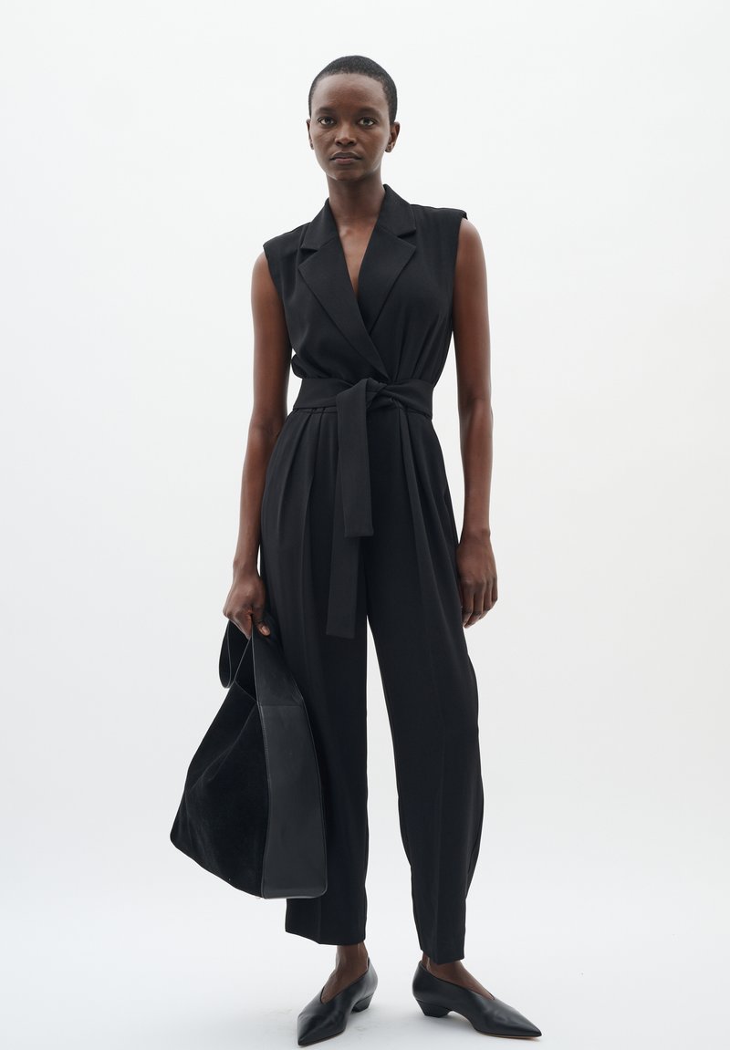Salopette sans manches noire avec un design croisé et une taille ceinturée, assortie à un sac noir et des ballerines à bout pointu sur un fond clair.