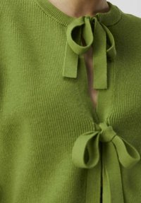 Haut en maille côtelée verte avec un col rond, une ouverture fendue à l'avant, et des nœuds décoratifs au niveau du col et de la taille.