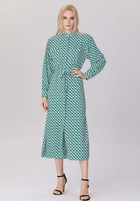 Femme debout portant une robe midi à motifs teal et blancs avec des manches longues et des talons à strap noir sur un fond uni.