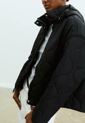 Personne portant une veste matelassée noire avec un col haut sur un haut gris clair et un pantalon blanc, debout devant un fond clair uni.