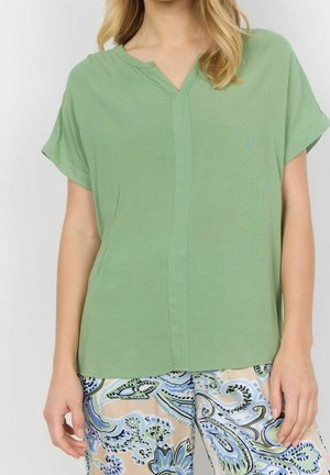 Grüne Bluse mit V-Ausschnitt, kurzen gerollten Ärmeln und glatter Textur; kombiniert mit gemusterten hellen Hosen in Blau und Creme.