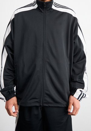 Homme portant une veste noire zippée intégralement avec des bandes blanches sur les manches et des poignets élastiques, debout devant un fond uni.