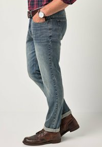 Vaqueros de denim azul claro con corte recto, dobladillos rematados y bolsillos frontales. Combinados con botas de cuero marrón y un reloj.