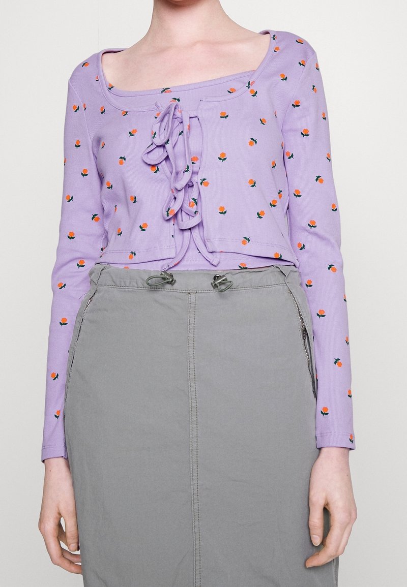 Femme portant un crop top lavande à manches longues avec de petits motifs floraux oranges et des liens à l'avant, associé à une jupe grise taille haute avec des poches zippées.
