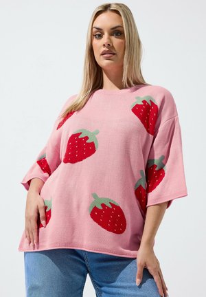 Lyshåret kvinde iført lyserød oversized sweater med store røde og grønne jordbærmønstre, kombineret med blå jeans, stående med hænderne hvilende.