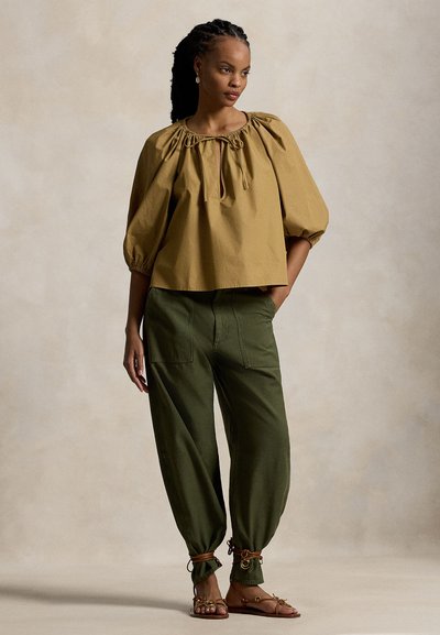 Μπλούζα χακί με φουσκωτά μανίκια και κορδόνι στο λαιμό, συνδυασμένη με ελιάς πράσινο, κωνικές παντελόνες με ελαστικές μανσέτες και τσέπες. Καφέ σανδάλια με λουράκια.