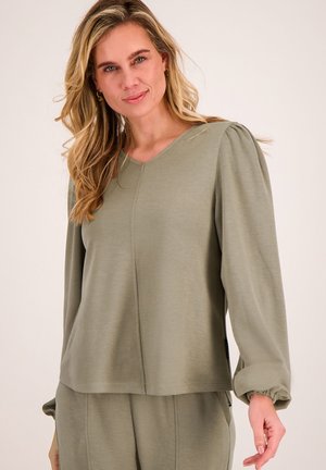 Blonde vrouw met loszittende olijfgroene top met lange mouwen en bijpassende broek, staand tegen een eenvoudige lichte achtergrond.