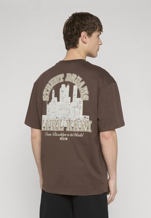 Ung mand set fra siden iført en brun oversize t-shirt med hvid bysilhuet og teksten "Street Dreams Karl Kani" på ryggen.