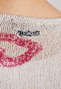 Gros plan d’un tissu beige tricoté avec une empreinte de lèvres rouges et une petite étiquette noire de la marque Desigual attachée.