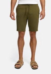 Hombre vestido con pantalones cortos de tiro concordante color verde oliva y sandalias beige, de pie frente a un fondo blanco liso.