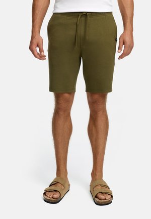 Mann iført olivengrønne snøreshorts og sandfarvede sandaler, stående foran en ensfarvet hvid baggrund.