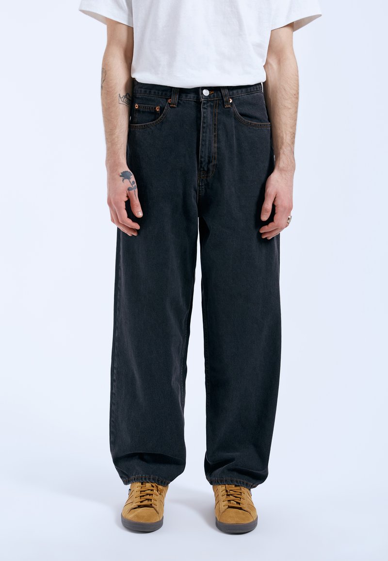 Black wide-leg jeans with five pockets, orange stitching, and a metal button closure. Worn with tan sneakers and a white t-shirt.