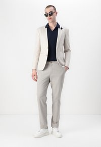 Beige blazer, marineblå teksturert skjorte, lys grå bukser og hvite joggesko. Blazeren har to frontlommer og knapplukking.