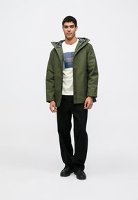 Casaco impermeável verde com capuz, combinado com uma t-shirt creme com uma estampa azul, calças pretas e sapatos pretos.