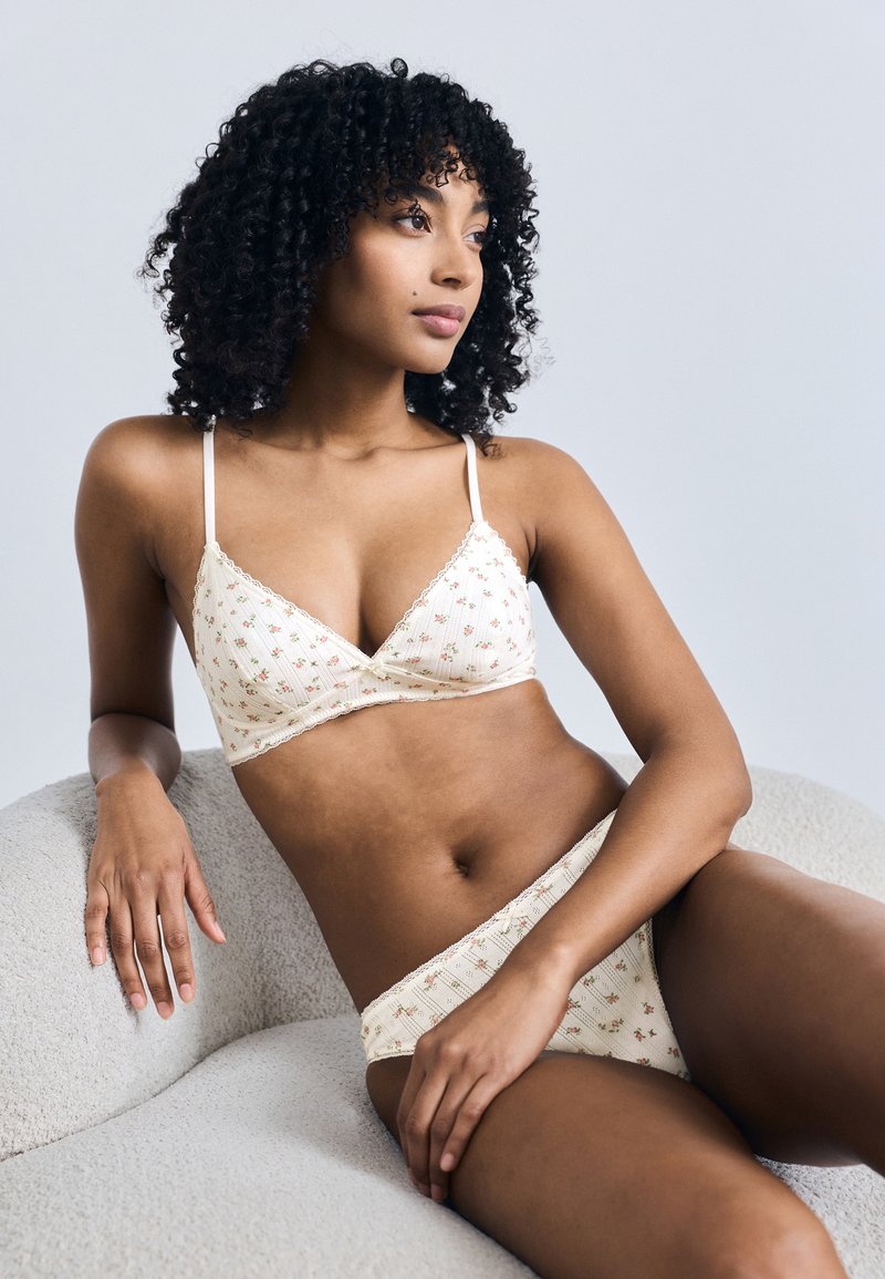 Γυναίκα με σγουρά μαλλιά που φοράει κρεμ σουτιέν με λουλουδάτο μοτίβο και εσώρουχα που ταιριάζουν, ξαπλωμένη σε μια καρέκλα με ανοιχτό γκρι υφή.