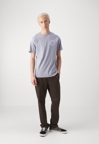 Helles Lavendelt-Shirt mit kurzen Ärmeln und rundem Ausschnitt, verziert mit einem weißen kreisförmigen Grafik. Kombiniert mit braunen Hosen und Schachbrett-Sneakers.