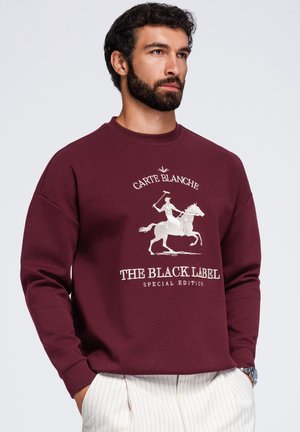 Homme portant un sweat-shirt bordeaux avec une broderie blanche d’un joueur de polo à cheval et le texte "Carte Blanche The Black Label Special Edition."
