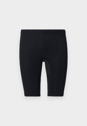 Mann står barfot, iført tettsittende svart kortermet genser med glidelås og matchende svarte shorts mot ensfarget hvit bakgrunn.