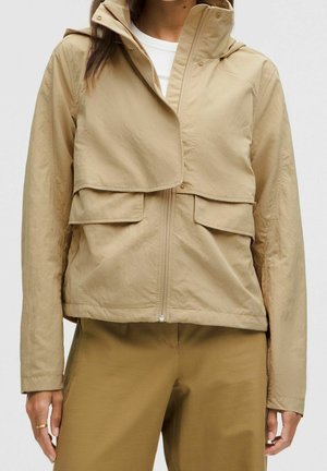 Femme portant une veste légère beige avec poches à rabat et un pantalon assorti, debout devant un fond uni.
