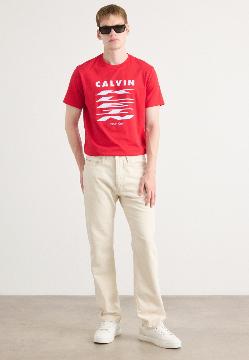 T-shirt en coton rouge avec un texte graphique blanc, associé à un pantalon en denim beige et des baskets blanches. Le design est simple et moderne.