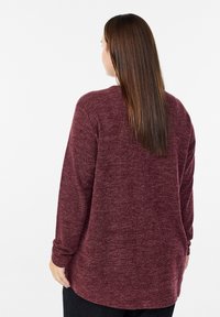 Długi rękaw, teksturowany top w kolorze bordowym, o luźnym kroju i okrągłym dole. Widok z tyłu ukazuje pojedynczą kieszeń na plecach.