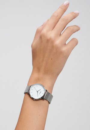 Sølvnetur armbåndsur på et håndled, med rund hvid urskive med sorte timeangivelser og visere, og et minimalistisk design.