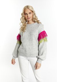 Lichtgrijze gebreide trui met lange mouwen, voorzien van heldere roze en groene franjes op de schouders. Een paar witte jeans.