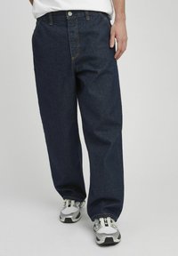 Jean en denim bleu foncé avec une coupe droite, présentant des coutures orange, des passants de ceinture et une fermeture à un seul bouton. Porté avec des baskets grises.