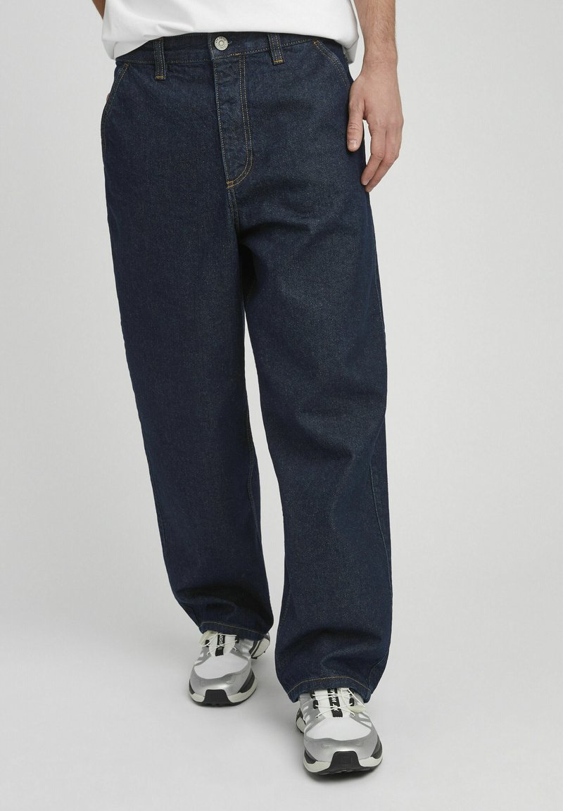 Jean en denim bleu foncé avec une coupe droite, présentant des coutures orange, des passants de ceinture et une fermeture à un seul bouton. Porté avec des baskets grises.