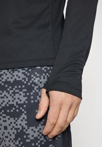 Maglietta sportiva nera a maniche lunghe con tessuto testurizzato e fori per i pollici, abbinata a pantaloni corti grigi con motivo a pixel.