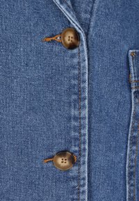 Farmerdzseki részlet, amely világoskék anyagból készült, egy fedeles zsebbel és két barna, négylyukú gombbal, narancssárga cérnával varrva.