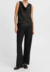 Top senza maniche in satin nero con colletto a cowl, abbinato a pantaloni neri texture morbidi. Indossato con scarpe scure. Design semplice, look monocromatico.