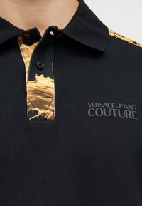Κοντινό πλάνο μαύρου πόλο πουκάμισου με μαύρα κουμπιά, εσωτερική πατιλέτα με χρυσό σχέδιο και λογότυπο "Versace Jeans Couture" στο στήθος.