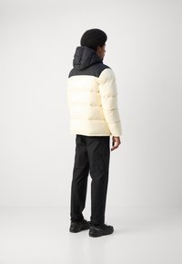 Giacca imbottita crema con spalle e cappuccio neri, caratterizzata da cuciture orizzontali. Indossata con pantaloni cargo neri e scarpe.