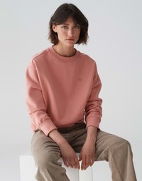 Felpa rosa con polsini a costine, collo rotondo e logo a cuore discreto sul petto, abbinata a pantaloni beige chiaro. Texture liscia.