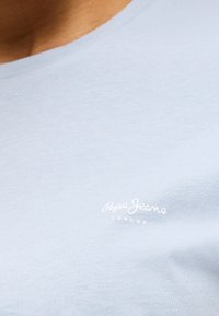 Detail svetlomodrej látky s bielym logom „Pepe Jeans London“ vytlačeným na oblasti hrude košele.