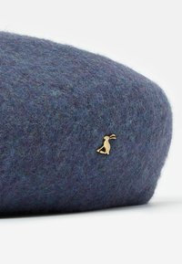 Plstená baretka v hlbokom modro-sivom odtieni, s textúrovaným povrchom a malým zlatým zajacom na boku.