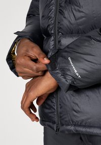 Schwarze, isolierte Jacke mit Reißverschluss, Klettverschlüssen an den Bündchen und "OMNI-HEAT"-Markierung. Der Stoff hat eine glatte, glänzende Textur.