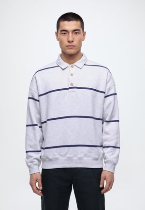 Un homme porte un sweat-shirt gris clair à col, à rayures horizontales bleu marine, et un pantalon noir sur un fond blanc uni.