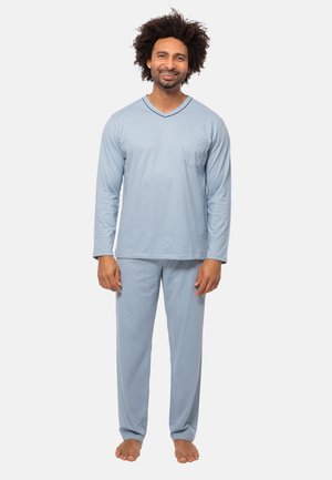 Mann steht barfuß, trägt ein hellblaues Langarmshirt und passende Hose, lächelt vor einem schlichten weißen Hintergrund.