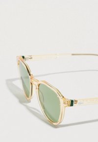 Transparante lichtbeige zonnebril met rund groene getinte glazen, rustend op een wit oppervlak.