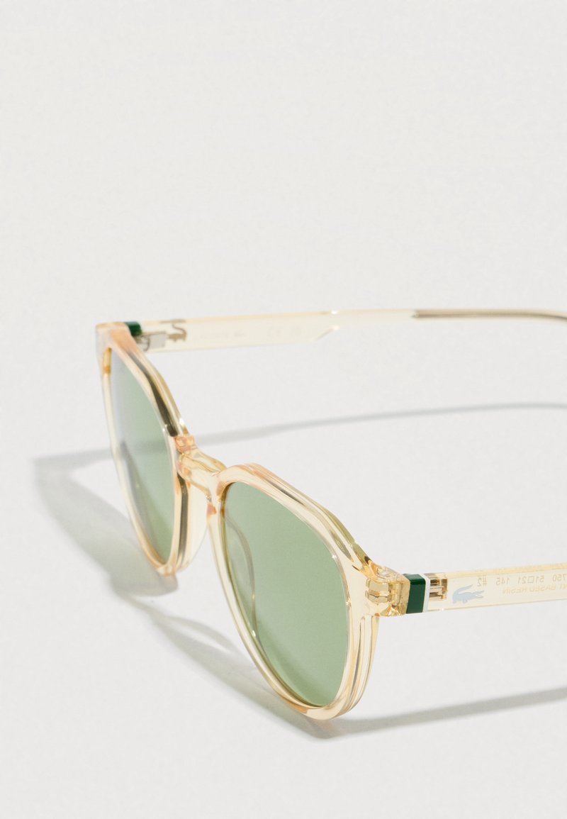 Lunettes de soleil à monture beige clair transparent avec des verres ronds teintés verts reposant sur une surface blanche.